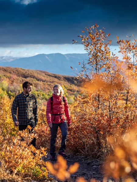 A man and a woman hiking in the mountains during the day, surrounded by fall foliage in shades of orange and brown.