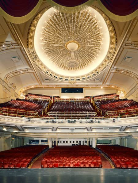 The view from the stage of a theater, showcasing ornately decorated ceilings and red theater chairs.
