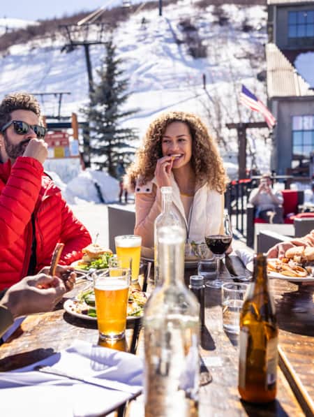 People at a ski resort, bundled up in winter clothing, eating and drinking at a wooden table outside.