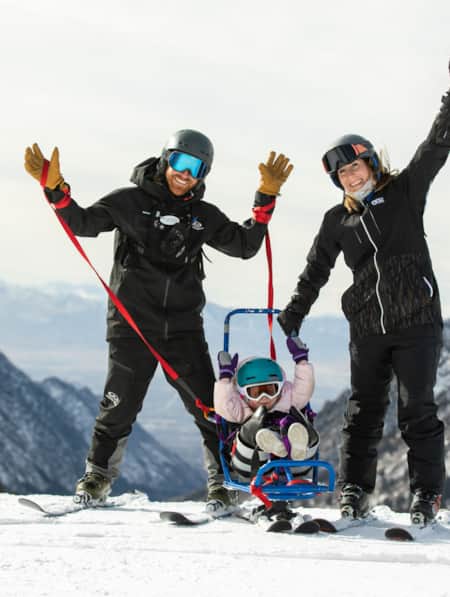 Two skiers smiling with their hands raised in the air, with a small child standing between them.